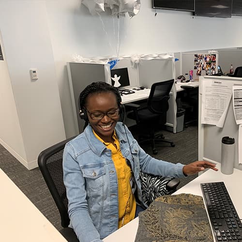 A smiling young woman wearing a denim jacket and yellow top is sitting at a desk in an office environment, with a computer and other office equipment visible in the background.