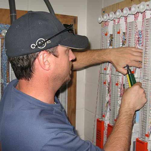 A man wearing a baseball cap is working on an electrical panel in a room with various household items and decorations visible in the background.