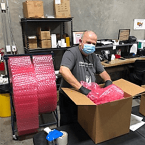A man wearing a face mask is packing boxes in an office setting with shelves and other boxes in the background.