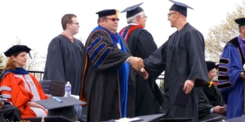 The image shows several individuals in graduation gowns and caps, shaking hands and conversing at what appears to be a graduation ceremony.