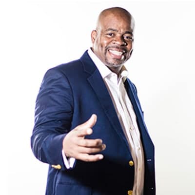 A smiling man in a navy blue suit stands against a plain white background, gesturing with his hands in a friendly and engaging manner.