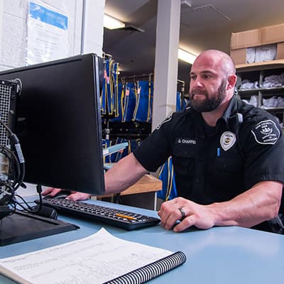 A uniformed police officer sits at a desk, working on a computer in an office setting with various supplies and equipment visible in the background.