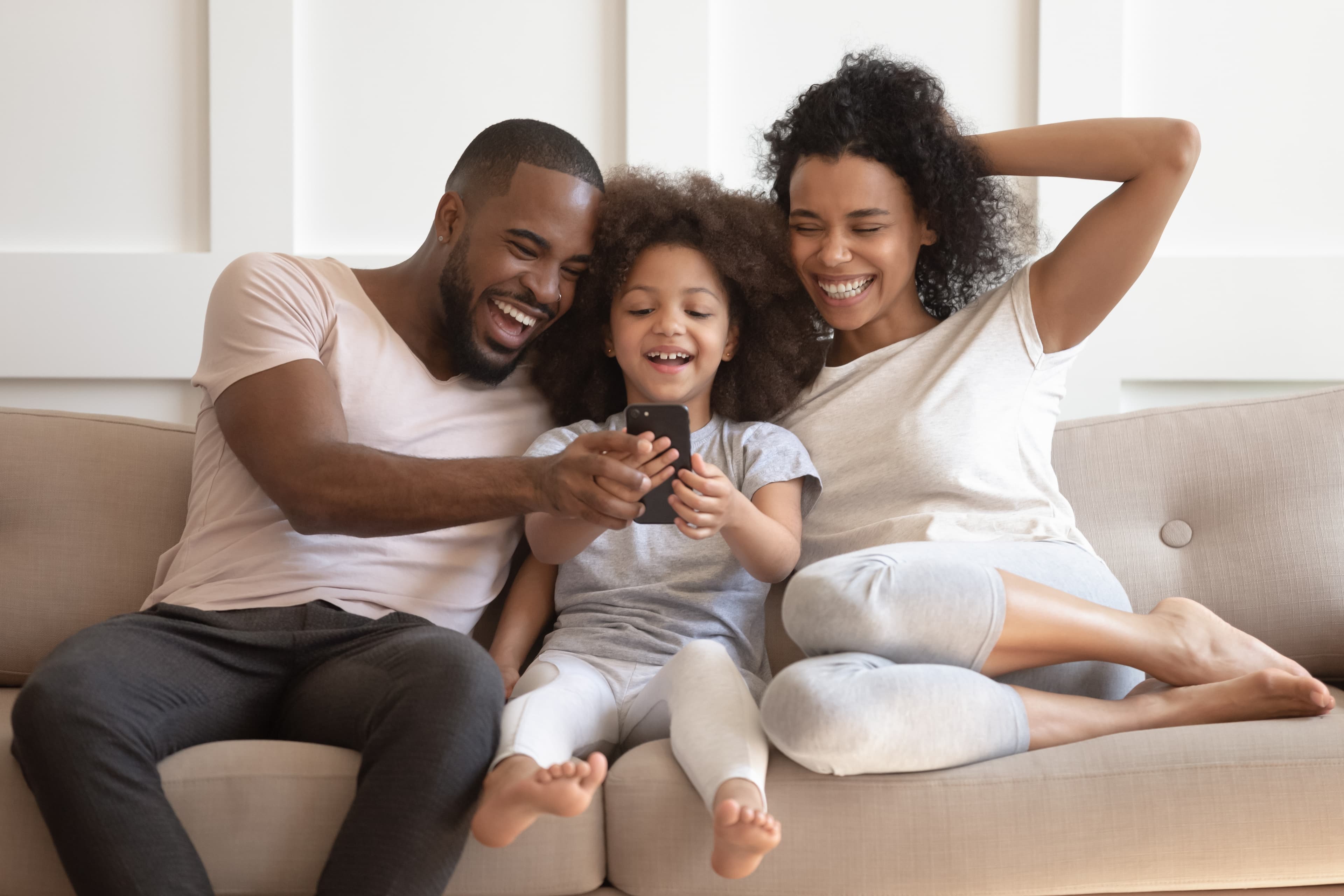 A happy family of three - a father, mother, and young daughter - sitting together on a couch, laughing and enjoying each other's company.
